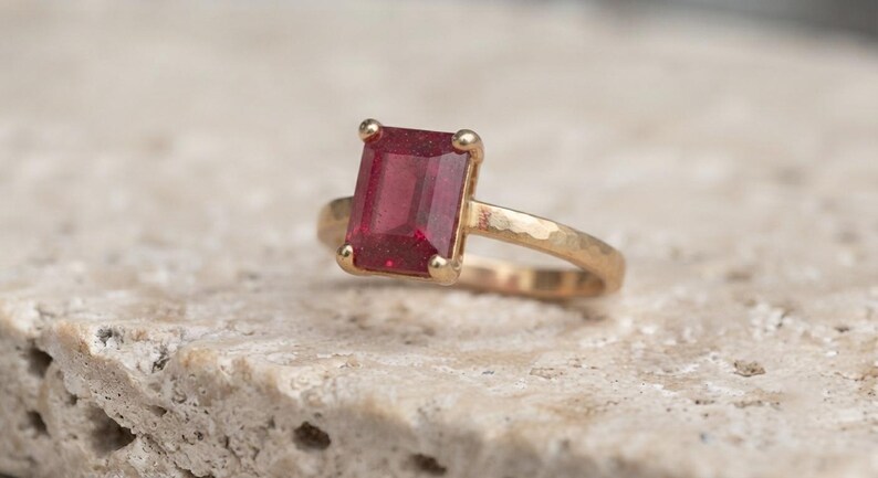 May include: A gold ring featuring a rectangular emerald-cut red gemstone set in a four-prong setting. The band of the ring has a distinct hammered texture, providing a matte finish to the metal surface that contrasts with the faceted red stone.