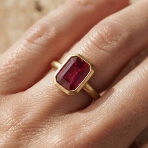 May include: A ring featuring a rectangular, emerald-cut deep red gemstone set in a smooth, matte-finish yellow gold bezel setting. The band is a simple, solid yellow gold design that complements the prominent, faceted red stone at the centre.
