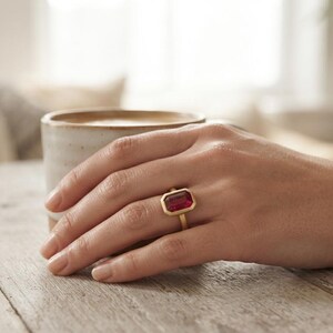 May include: A gold ring featuring a large, rectangular, deep red gemstone set in a smooth metal bezel. The ring is worn on a finger, showcasing the vibrant colour of the stone and the polished finish of the gold band against the skin.