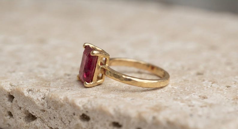 May include: A gold ring featuring a rectangular, emerald-cut red gemstone set in a four-prong setting. The ring band has a distinct hammered texture, providing a reflective surface that complements the deep red colour of the central stone.