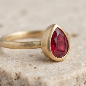 Può includere: Un anello in oro con una pietra preziosa rossa sfaccettata a forma di pera incastonata in una lunetta liscia. La fascia dell'anello presenta una finitura martellata e testurizzata che contrasta con l'incastonatura lucida della pietra.
