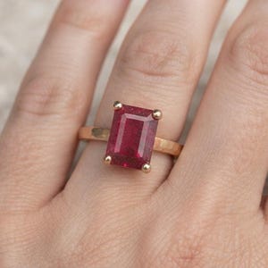 May include: A gold ring featuring a rectangular, emerald-cut deep red gemstone set in a four-prong setting. The band of the ring has a textured, hammered finish, providing a distinct surface appearance compared to a smooth, polished metal band.
