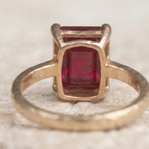 May include: A gold ring featuring a rectangular, emerald-cut deep red gemstone set in a four-prong setting. The band of the ring has a textured, hammered finish, and the metal appears to have a warm, yellow-gold tone throughout the entire piece.