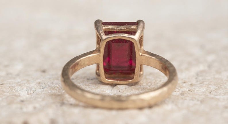 May include: A gold ring featuring a rectangular, emerald-cut deep red gemstone set in a four-prong setting. The band of the ring has a textured, hammered finish, and the metal appears to have a warm, yellow-gold tone throughout the entire piece.