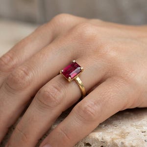 May include: A gold ring featuring a rectangular, emerald-cut deep pink gemstone set in a four-prong setting. The band of the ring has a textured, hammered finish, providing a distinct surface appearance compared to a smooth, polished metal band.
