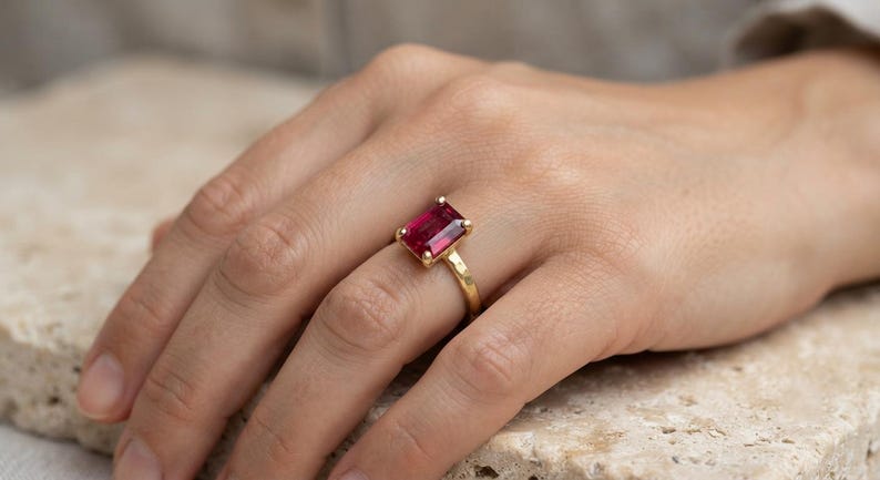 May include: A gold ring featuring a rectangular, emerald-cut deep pink gemstone set in a four-prong setting. The band of the ring has a textured, hammered finish, providing a distinct surface appearance compared to a smooth, polished metal band.