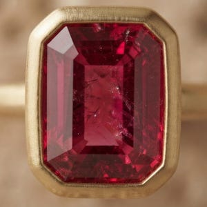 May include: A ring featuring a large, rectangular, faceted deep pink gemstone set in a smooth, brushed yellow gold bezel setting. The stone displays internal inclusions and a vibrant, saturated colour, while the gold band has a matte finish.