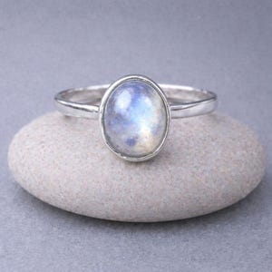 May include: A silver ring featuring an oval moonstone with iridescent qualities. The gemstone presents a blend of blue, white, and yellow tones. The ring is displayed on a smooth, grey stone, highlighting the jewellery's design and colour.