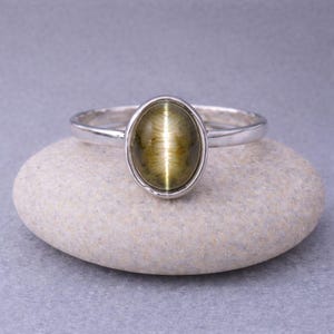 Op de afbeelding: Zilveren ring met een ovale, olijfgroene edelsteen. De edelsteen vertoont een chatoyance-effect, met een enkele lichtband. De ring ligt op een lichtgrijze steen. De band is effen zilver.