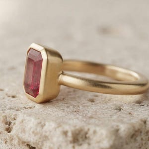 May include: A gold ring featuring a rectangular, emerald-cut red gemstone set in a smooth, bezel-style mounting. The band has a matte, brushed finish, and the gemstone is positioned vertically, showcasing its deep red colour and faceted surface.