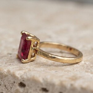 May include: A gold ring featuring a rectangular, deep pink gemstone set in a four-prong setting. The band of the ring has a distinct hammered, textured finish, providing a contrast to the smooth, faceted surface of the central stone.