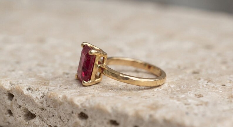 May include: A gold ring featuring a rectangular, deep pink gemstone set in a four-prong setting. The band of the ring has a distinct hammered, textured finish, providing a contrast to the smooth, faceted surface of the central stone.
