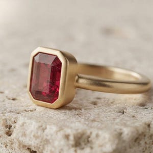 May include: A gold ring featuring a rectangular, emerald-cut deep red gemstone set in a smooth, matte-finish gold bezel. The band is solid gold with a rounded profile, showcasing a clean, simple design that highlights the vibrant red stone.