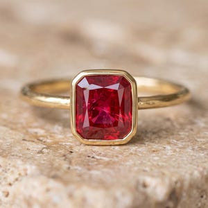 May include: A gold ring featuring a rectangular, faceted, deep red gemstone set in a smooth, polished gold bezel. The band of the ring is thin, rounded, and crafted from a matching yellow gold metal, creating a clean and simple design.