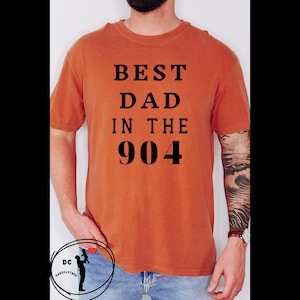 May include: Burnt orange t-shirt with the text "BEST DAD IN THE 904" in black. The shirt has short sleeves and a crew neck. A small logo is visible at the bottom left. The person is wearing a watch with a red band.