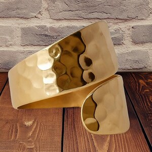 May include: A gold cuff bracelet with a hammered finish. The bracelet's design is open, with one end overlapping the other. The surface reflects light, creating highlights and shadows. The jewellery is displayed on a wooden surface.