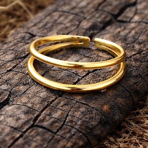 Könnte beinhalten: Ein goldfarbener Doppelring mit offenem Design. Der Ring wird auf einer verwitterten, dunkelbraunen Holzoberfläche mit sichtbarer Maserung und Textur präsentiert. Die Ringbänder sind glatt und glänzend und reflektieren das Licht.
