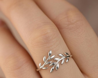 May include: A silver-colored ring featuring a delicate leaf design. The ring has two branches of leaves that curve around the finger. The leaves have a smooth, polished finish.