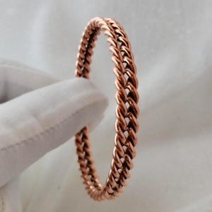 May include: A copper-colored bracelet with a braided chain design. The bracelet is a continuous loop, showcasing a detailed weave pattern. The warm, metallic hue of the bracelet is highlighted against a soft, white background.