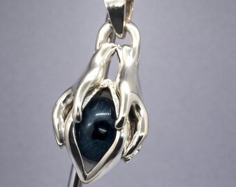 Puede incluir: Un colgante de plata con dos manos estilizadas sosteniendo una gema azul oscuro. El colgante cuelga de una cadena dorada. Las manos están intrincadamente detalladas y la gema tiene una superficie pulida. El diseño es artístico y simbólico.