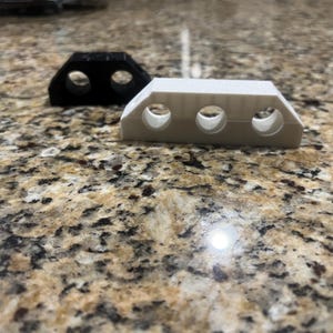 May include: Two plastic components, one black and one off-white, each with two or three circular holes. The objects are rectangular with angled sides. The items are on a speckled, brown and tan surface.