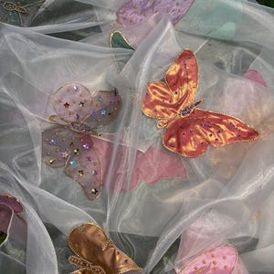 May include: Sheer white fabric adorned with multiple embroidered butterflies in shades of pink, purple, and orange. The butterflies have gold trim and are embellished with small, colorful beads, creating a delicate and whimsical design.