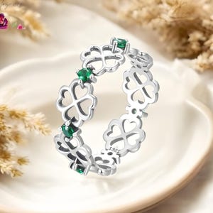 Puede incluir: Un anillo de plata con diseño de trébol, adornado con piedras preciosas verdes. El anillo se muestra en un plato blanco, con el nombre de la marca "ELAROSA". El fondo incluye plantas secas.