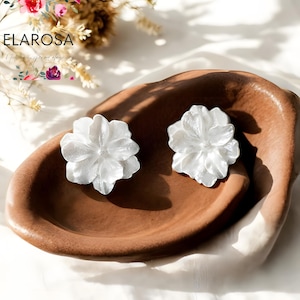 Puede incluir: Dos pendientes de botón blancos con forma de flor, cada uno con un diseño de pétalos texturizados, presentados sobre un plato ovalado marrón. Los pendientes están sobre un fondo blanco con decoraciones florales. La marca "ELAROSA" es visible.