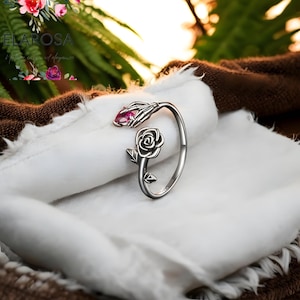 Può includere: Anello argentato con una mano scheletrica che tiene una gemma rosa e una rosa. L'anello è posto su un tessuto bianco, con uno sfondo marrone e verde. Il design dell'anello è intricato e dettagliato.