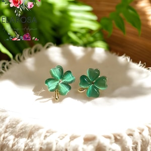 Puede incluir: Un par de pendientes verdes con forma de trébol con tallos dorados. Los pendientes están colocados sobre una superficie blanca, con un fondo borroso de follaje verde. Son una joya.
