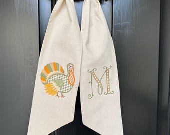 May include: A beige Thanksgiving door hanger with a colorful embroidered turkey and the letter "M". The door hanger is hanging on a black door with a green wreath in the background. The door hanger is made of a soft, linen-like material.