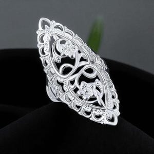 Puede incluir: Un anillo plateado con un diseño alargado y ornamentado. El anillo presenta intrincados patrones florales y de filigrana, con un motivo floral central. El anillo se encuentra sobre un fondo negro.