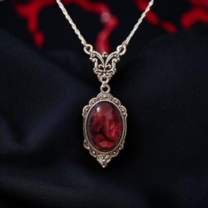 Può includere: Una collana argentata con un pendente ovale rosso sangue. Il pendente è incorniciato da una montatura ornata in stile filigrana. La catena è delicata e lo sfondo è un tessuto scuro e strutturato, creando un contrasto sorprendente.