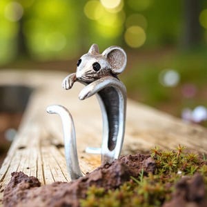 May include: A silver-colored ring featuring a detailed mouse design. The mouse has small black eyes and is perched on the ring's band, with its paws seemingly holding onto the ring. The ring is set against a natural outdoor background.