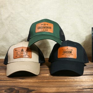 May include: Three baseball caps with mesh backs. One is green with "GROOMSMAN" on a leather patch, another is tan with initials and a date, and the third is black with "GROOM" on a leather patch.