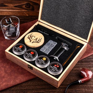 Puede incluir: Un juego de regalo de madera para entusiastas del whisky, que presenta una caja de madera con varios accesorios. Incluye piedras de whisky, virutas de madera en latas, un soplete, una cuchara y una pipa. La caja tiene el texto "Whiskey Stones" y un logotipo "CAB".