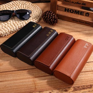 May include: Four leather eyeglass cases in black, dark brown, medium brown, and light brown, each with initials in gold. Sunglasses rest on a woven mat. The cases are on a wooden surface. The cases are approximately 16 cm long.