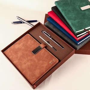 May include: A brown leather journal with a metal clasp and the initials "KY" is displayed in a gift box, alongside two pens. Other journals in various colours, including green, red, and blue, are visible in the background.