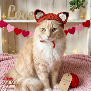 Puede incluir: Un gato naranja y esponjoso con un gorro de zorro naranja tejido con detalles en negro y blanco. El gato está sentado sobre una manta rosa, con una bola de hilo roja y una etiqueta que dice "Handmade with Love". La imagen también incluye el texto "4 Easy Cat Hat Patterns (PDF Download)".