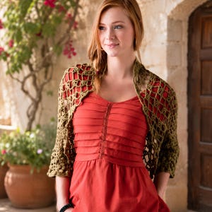 May include: A red dress with a pleated bodice is paired with a crocheted olive green shawl. The shawl has an open, net-like design. The background includes a stone wall, a wooden door, and potted plants with pink flowers.