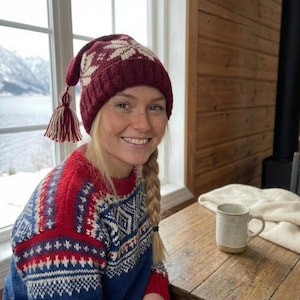 Könnte beinhalten: Eine bordeauxfarbene Strickmütze mit weißem Schneeflockenmuster und Quastendetails. Die Mütze wird mit einem rot-weiß-blau gemusterten Pullover getragen. Der Pullover hat ein geometrisches Design.