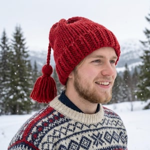 Men's or unisex chunky red winter beanie modeled outdoors. Close up of the heavy knit texture and large yarn tassel side detail in a winter snow setting.