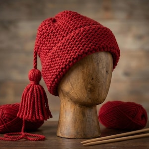Studio shot of a thick red yarn beanie with tassel on a hat display stand, set against a rustic wooden background with knitting supplies.