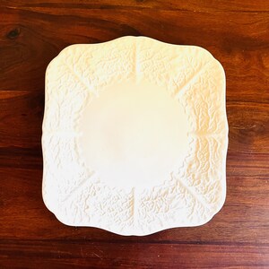 May include: Off-white, square-shaped ceramic plate with a cabbage leaf design. The plate has a slightly raised, textured surface and a smooth, central area for food. The plate is set on a dark wooden surface.