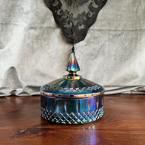 May include: An iridescent glass jar with a lid. The jar is round with a diamond pattern on the sides and a ridged top. The lid has a pointed finial. The colors shift from blue to purple to green. The jar sits on a wooden surface.
