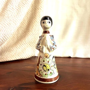 May include: A ceramic figurine of a person with black hair and a white dress adorned with floral patterns in green, yellow, and blue. The figurine has a brown base and a brown collar. The hands are clasped in front. The figurine is standing on a wooden surface.
