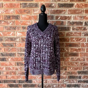 May include: A V-neck sweater in a textured knit pattern, displayed on a black mannequin. The sweater features a mix of dark purple and white threads, creating a mottled effect. The sleeves are long, and the overall style is casual.