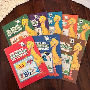 May include: A collection of vintage "Big Bird's Sesame Street Dictionary" books. Each book features Big Bird on the cover, with different colored covers and volume numbers. The books are arranged on a dark wooden surface.