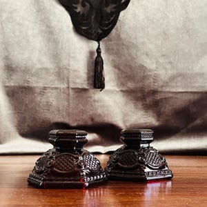 May include: Two dark red glass candle holders with ornate, geometric designs. The candle holders are square-shaped with a detailed pattern on the sides and a flat top. They sit on a wooden surface against a textured background.
