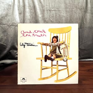 May include: Album cover for "And That's the Truth" by Lily Tomlin. The cover features a photo of a young girl sitting in a yellow rocking chair, holding a doll. The album title and artist's signature are in pink and black.